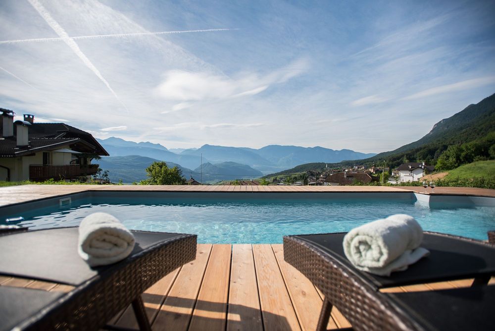 Ein Swimmingpool auf einer Holzterrasse mit Liegen und Handtüchern, dahinter ein Dorf und bewaldete Berge unter blauem Himmel.