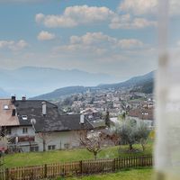 Blick über Dächer und einen Garten auf ein Dorf in einem Bergtal; rechts verdeckt ein unscharfer heller Vorhang teilweise die Aussicht.