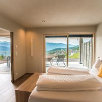 Modernes Schlafzimmer mit weiß bezogenem Bett und gelben Kissen; durch große Glasschiebetüren sieht man einen Balkon und eine Berglandschaft.