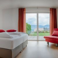 Helles Schlafzimmer mit großem weiß bezogenem Bett, roten Akzenten und rotem Sessel; durch die Glastür sind Garten und Berge zu sehen.