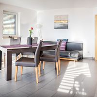 Heller Wohn- und Essbereich mit Esstisch und vier Stühlen, grauem Sofa, offener Küche und großen Glastüren zum Balkon.