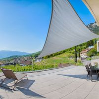 Sonnige Terrasse mit großem Sonnensegel, Liegestühlen und Esstisch; dahinter liegen Weinberge, ein Dorf und Berge.
