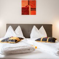 Ein ordentlich gemachtes Doppelbett mit weißen Kissen und gelb-schwarzen Zierkissen steht vor einem grauen Kopfteil.