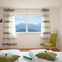 Helles Schlafzimmer mit einem Bett, grünem Sessel und einem Fenster mit Blick auf Berge.