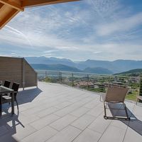 Große geflieste Terrasse mit Esstisch, Weinflasche und Liegestühlen; dahinter liegen ein Dorf und bewaldete Berge unter blauem Himmel.