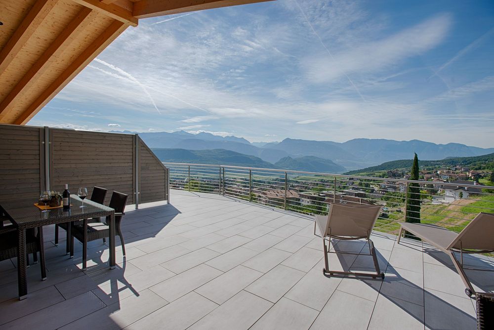 Große geflieste Terrasse mit Esstisch, Weinflasche und Liegestühlen; dahinter liegen ein Dorf und bewaldete Berge unter blauem Himmel.