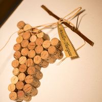Eine aus Weinkorken geformte Traube hängt an einem Gestell aus Zweigen; daran ist ein beschrifteter Anhänger befestigt.