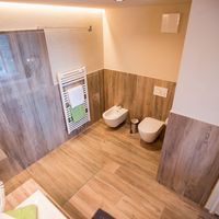 Modernes Bad mit bodengleicher Glasdusche, Bidet, WC und Handtuchheizkörper; Boden und Wände sind mit Holzoptikfliesen gestaltet.