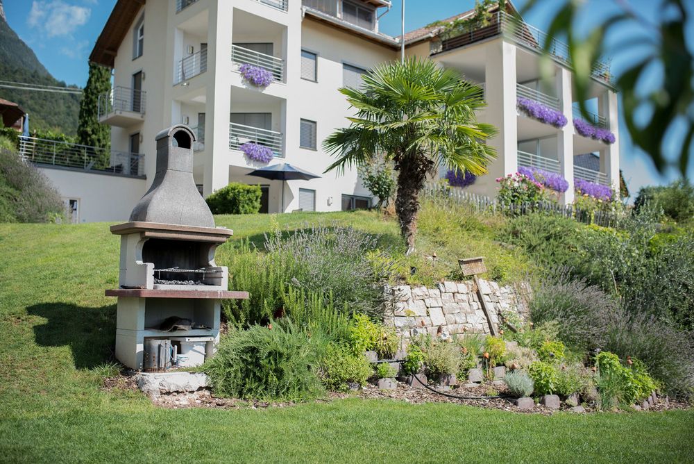 Ein weißes Mehrfamilienhaus mit Balkonen und violetten Blumen liegt hinter einem gepflegten Garten mit Palmenbaum und steinernem Gartengrill.