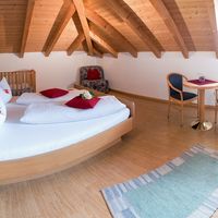 Helles Dachzimmer mit Holzbalkendecke, großem Bett, Kinderbett, Sessel und Sitzecke am Fenster.