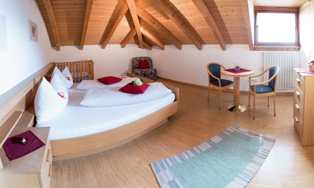 Helles Dachzimmer mit Holzbalkendecke, großem Bett, Kinderbett, Sessel und Sitzecke am Fenster.