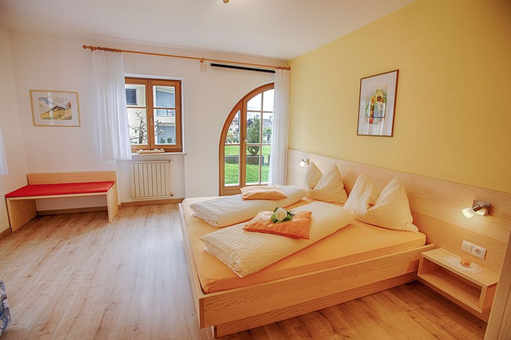 Helles Schlafzimmer mit großem Holzbett, weißer Bettwäsche, gelben Wänden und einer Rundbogentür zum Garten.