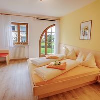 Helles Schlafzimmer mit großem Holzbett, weißer Bettwäsche, gelben Wänden und einer Rundbogentür zum Garten.