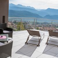 Eine sonnige Terrasse mit Loungemöbeln bietet einen Blick über ein Dorf auf bewaldete Berge.