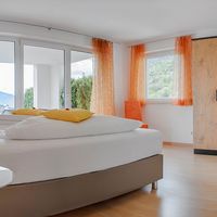 Helles Schlafzimmer mit großem Bett, orangefarbenen Vorhängen und einem Holzkleiderschrank; durch die Fenster ist eine grüne Landschaft zu sehen.