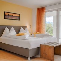 Helles Schlafzimmer mit großem weiß bezogenem Bett, gelber Wand und Glastür zur Terrasse mit Bergblick.