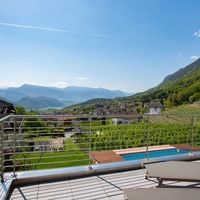 Blick von einer Terrasse mit Pool über ein Dorf und Weinberge in einem grünen Bergtal.