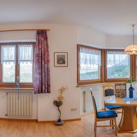 Helles Wohnzimmer mit blauem Sofa, Fernseher und Esstisch vor großen Fenstern mit Bergblick.