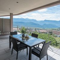 Überdachte Terrasse mit Gartentisch, Korbsesseln und Sonnenliegen; dahinter liegen ein Dorf und ein weites Bergpanorama.
