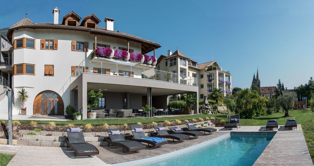 Modernes Hotelgebäude mit blumengeschmückten Balkonen, Liegestühlen und einem Außenpool; dahinter ist ein Kirchturm zu sehen.