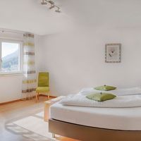 Helles Schlafzimmer mit großem weiß bezogenem Bett, grünem Stuhl und Fenster mit Blick auf eine Berglandschaft.