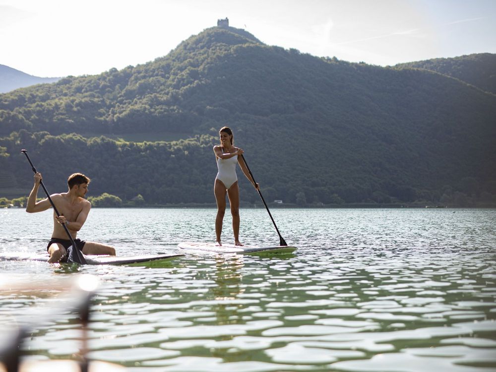 Eine Frau steht mit einem Paddel auf einem SUP-Board, während ein Mann auf einem zweiten Board sitzt; dahinter liegen ein See und bewaldete Berge mit einer Burg auf dem Gipfel.