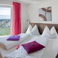 Helles Schlafzimmer mit zwei weißen Betten, violetten Kissen und pinken Vorhängen an einer Balkontür mit Blick auf Häuser und Berge.