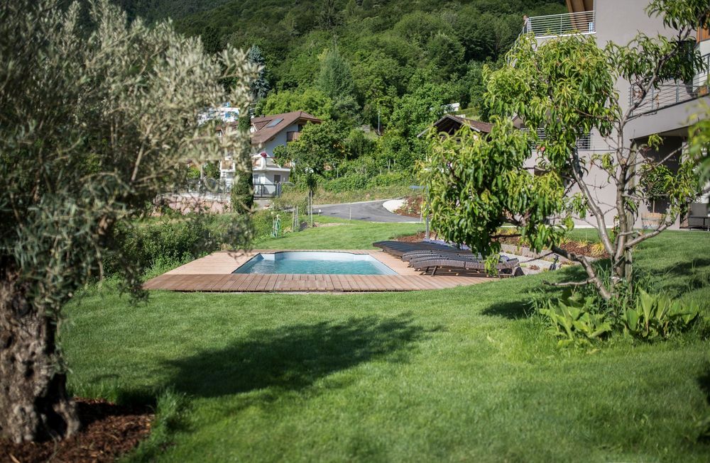 Ein kleiner Pool mit Holzumrandung liegt auf einer grünen Rasenfläche; daneben stehen Liegestühle, dahinter Häuser und ein bewaldeter Hang.