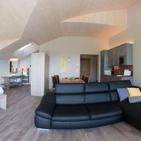 Helles, offenes Dachgeschosszimmer mit Doppelbett, schwarzer Sofaecke, Esstisch und Küchenzeile unter einer markanten Holzdecke.