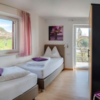 Helles Gästezimmer mit zwei Einzelbetten, violetten Kissen und Blick auf Weinberge sowie einen Balkon.