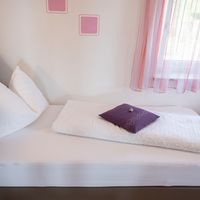 Ein weiß bezogenes Bett mit weißen und violetten Kissen steht neben einem Fenster mit rosafarbenen Vorhängen.