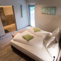 Modernes Schlafzimmer mit großem Doppelbett, weißer Bettwäsche und grünen Kissen; daneben führen Türen zu einem Bad und einem Balkon.