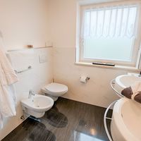 Helles Badezimmer mit Badewanne, Waschbecken, Toilette, Bidet und weißen Bademänteln am Fenster.