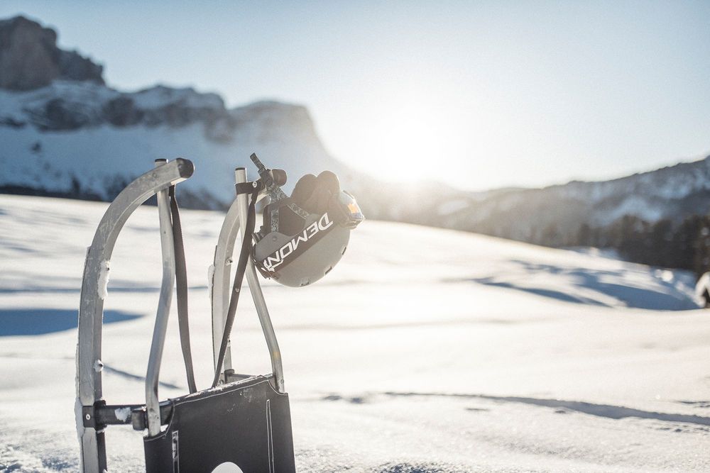 Ein Skihelm hängt an einer Metallhalterung auf einer verschneiten Skipiste vor schneebedeckten Bergen.