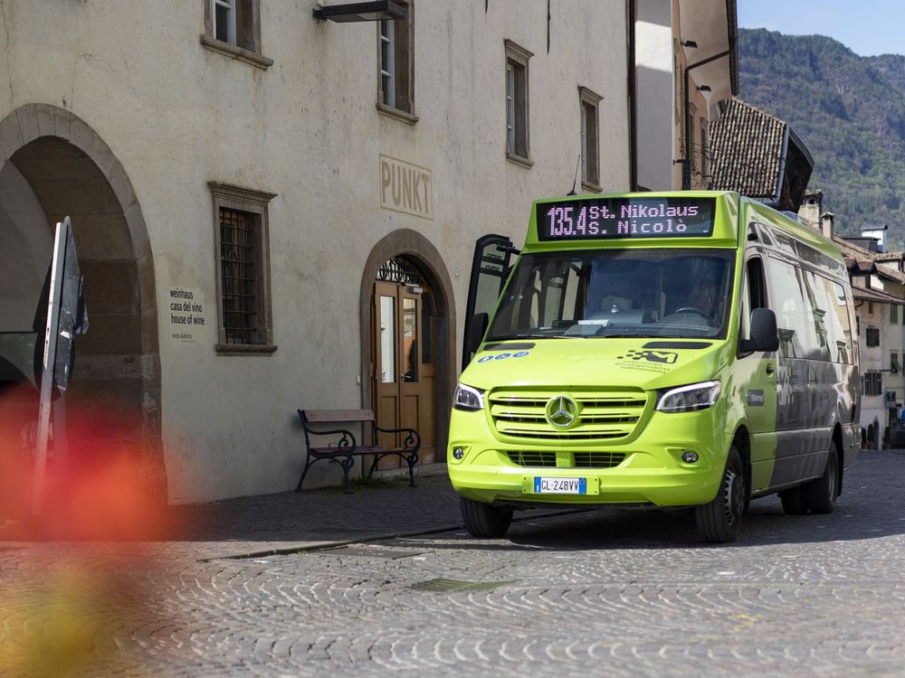 Ein hellgrüner Mercedes-Minibus steht auf einer Kopfsteinpflasterstraße vor einem historischen Gebäude in einem Bergort.