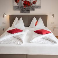 Ein ordentlich gemachtes Doppelbett mit weißer Bettwäsche und roten Kissen steht vor einem braunen Kopfteil; darüber hängt ein mehrteiliges Bild mit roten Blumen.