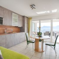 Heller Wohnbereich mit grauer Einbauküche, rundem Esstisch und großen Glastüren zu einem Balkon mit Bergblick.
