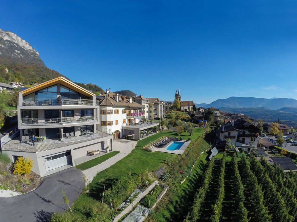 Bergdorf mit modernen Häusern, einem Pool und einer Kirche vor bewaldeten Bergen und blauem Himmel.