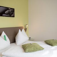 Ein weiß bezogenes Bett mit weißen und grünen Kissen steht vor einer hellgrünen Wand mit gerahmtem Steinmotiv.