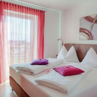 Helles Schlafzimmer mit zwei Einzelbetten, weißen Kissen und pinken Akzenten vor einem Fenster mit roten Vorhängen.