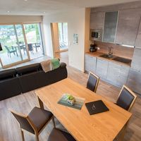 Heller Wohn- und Essbereich mit grauer Küche, schwarzem Sofa und großem Esstisch; durch die Glasschiebetüren ist eine Terrasse mit Sitzmöbeln zu sehen.