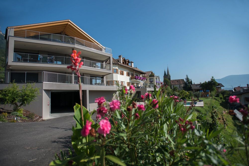Modernes Wohnhaus mit Balkonen und Holzdach steht neben älteren Häusern und einer Kirche; pinke Blumen wachsen im Vordergrund.