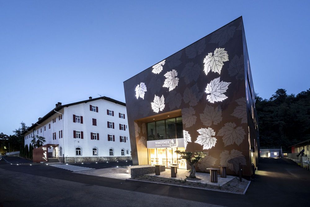 Eine moderne Vinothek mit beleuchteten Blattmustern an der dunklen Fassade steht neben einem traditionellen weißen Gebäude bei Dämmerung.