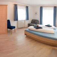 Helles Schlafzimmer mit großem Holzbett, Kleiderschrank, blauem Vorhang und kleiner Couch.