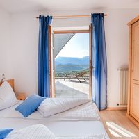 Helles Schlafzimmer mit weiß-blau bezogenen Betten, offenem Balkontürfenster und Blick auf Berge.
