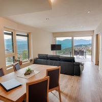 Heller Wohn- und Essbereich mit schwarzem Sofa, Holztisch und offener Küche; durch große Fenster und die Balkontür sind Berge und ein Ort zu sehen.