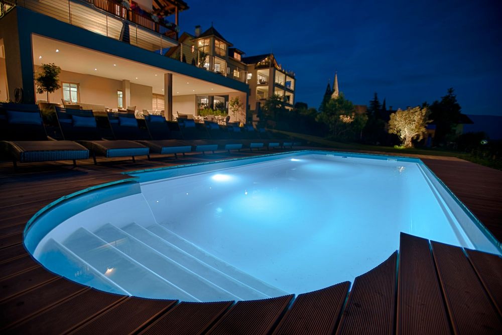 Ein beleuchteter Außenpool mit Holzterrasse und Liegen vor einem mehrstöckigen Hotel in der Abenddämmerung.