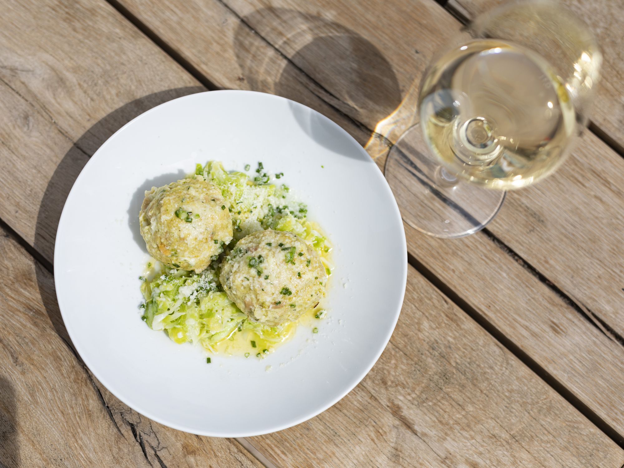 Zwei Kräuterknödel mit grünem Gemüse und geriebenem Käse auf einem weißen Teller; daneben steht ein Glas Weißwein auf einem Holztisch.