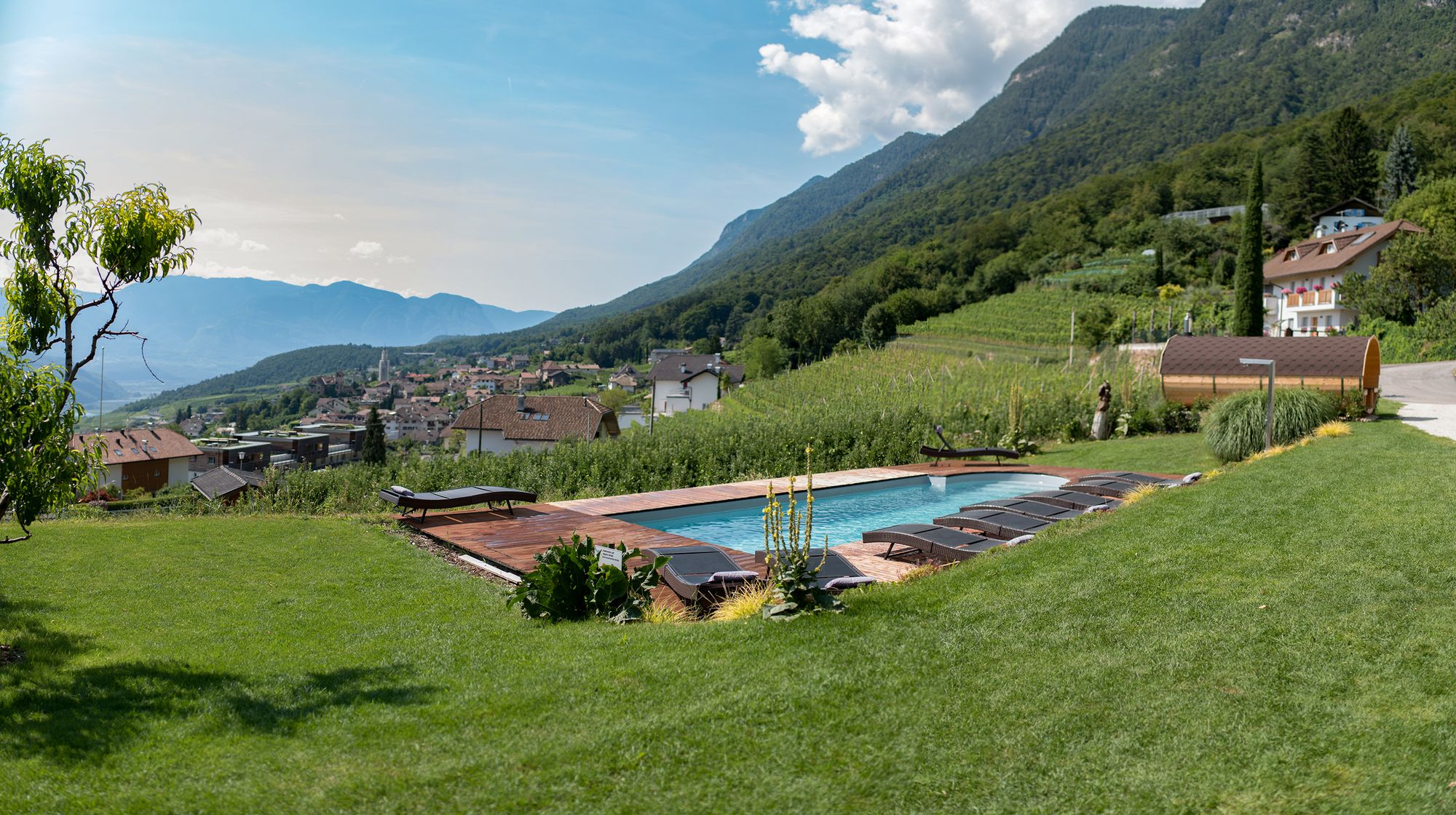 Ein Außenpool mit Liegestühlen liegt auf einer grünen Terrasse oberhalb eines Dorfes, umgeben von Weinbergen und bewaldeten Bergen.