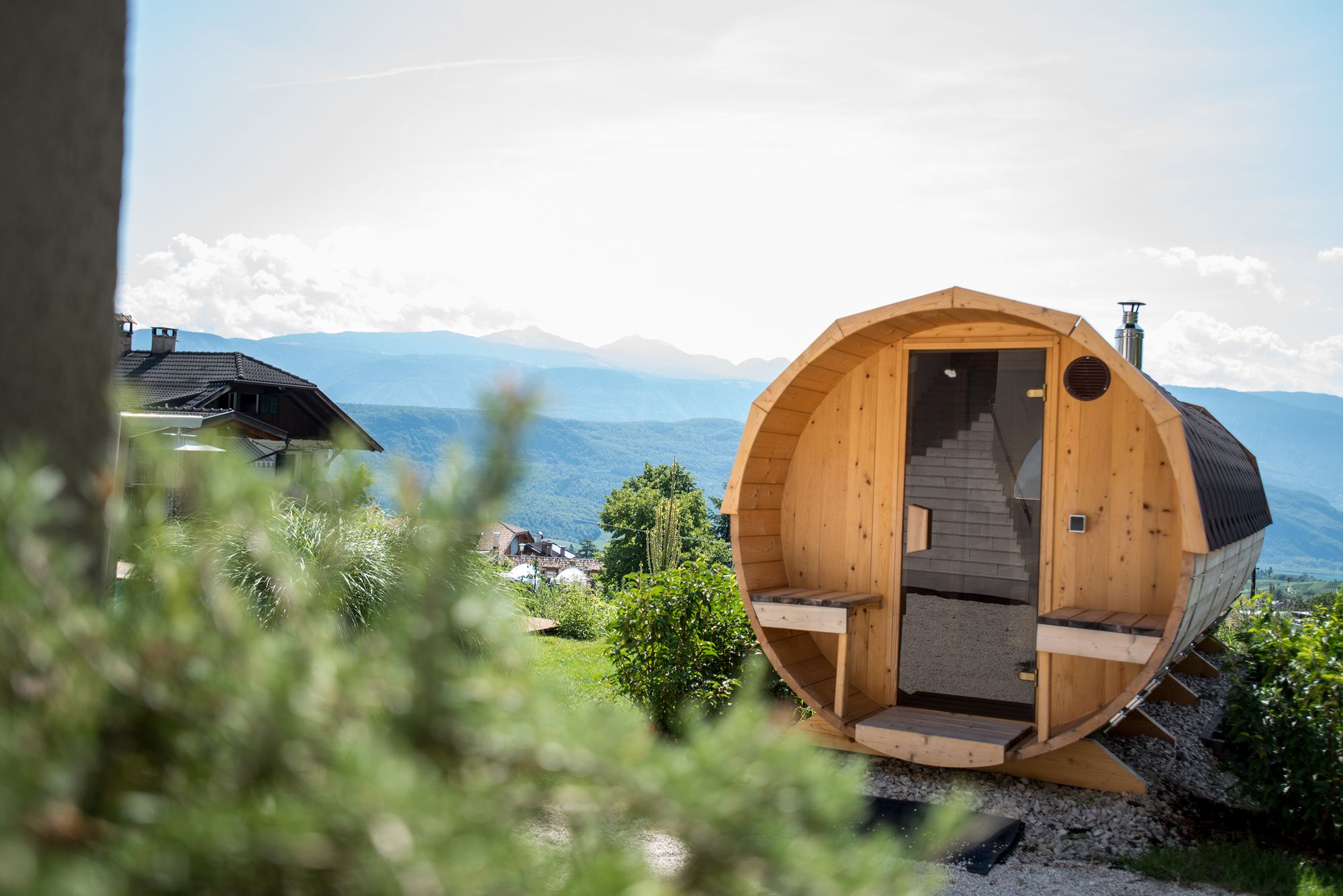 Eine tonnenförmige Holzsauna steht auf einer Terrasse vor einer Berglandschaft.